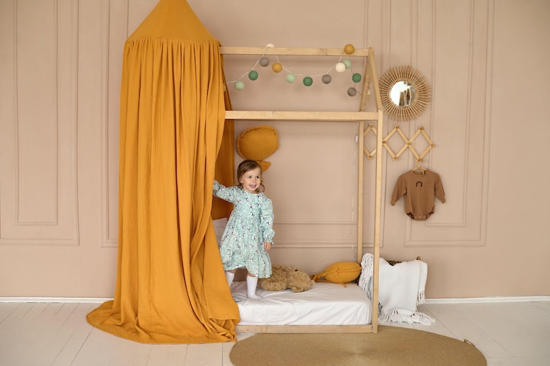 May include: A young girl in a blue and white floral dress stands inside a wooden playhouse with a yellow canopy. The playhouse has a white mattress and a stuffed animal on it. The playhouse is decorated with a string of lights and a wooden mirror.