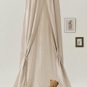 Pu&ograve; includere: Una tenda a baldacchino in gingham beige e bianco per la camera da letto di un bambino. La tenda &egrave; realizzata in tessuto morbido e ha una parte superiore arricciata. Un orsacchiotto di peluche marrone &egrave; seduto sul pavimento davanti alla tenda.