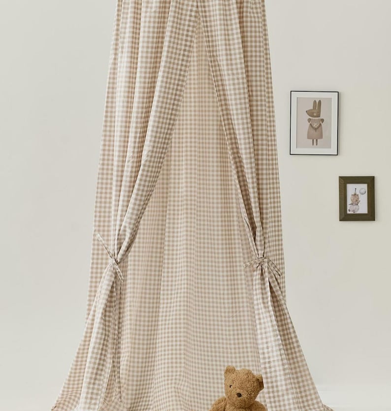 Pu&ograve; includere: Una tenda a baldacchino in gingham beige e bianco per la camera da letto di un bambino. La tenda &egrave; realizzata in tessuto morbido e ha una parte superiore arricciata. Un orsacchiotto di peluche marrone &egrave; seduto sul pavimento davanti alla tenda.