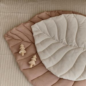 Pode incluir: Dois tapetes de brincar em forma de folha em rosa empoeirado e creme, com detalhes acolchoados. Duas pequenas figuras de árvore de madeira são colocadas no tapete rosa empoeirado. Os tapetes são projetados para bebês e crianças pequenas.