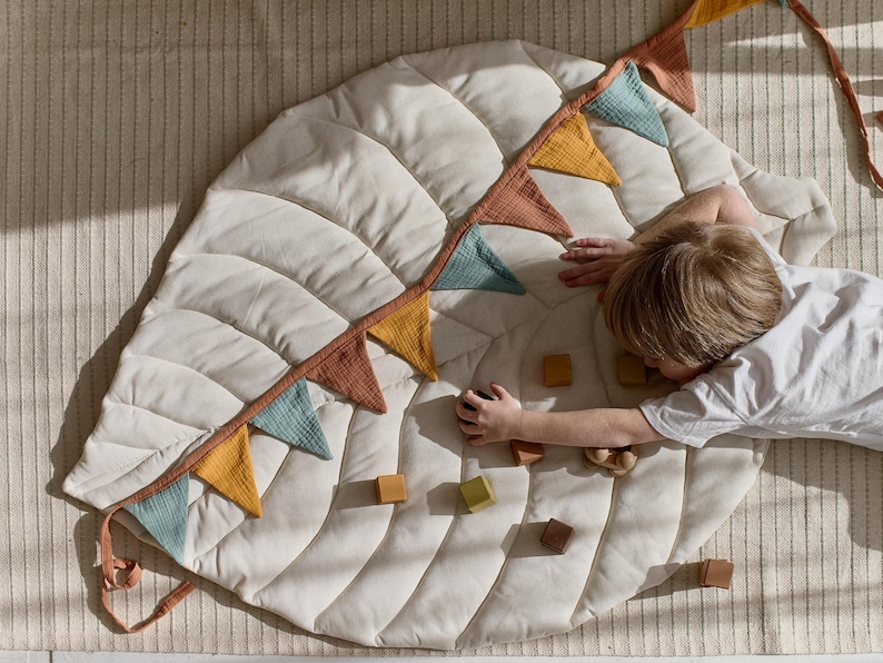 May include: A white quilted play mat shaped like a leaf with a colorful fabric bunting around the edge. A child is playing with wooden blocks on the mat.