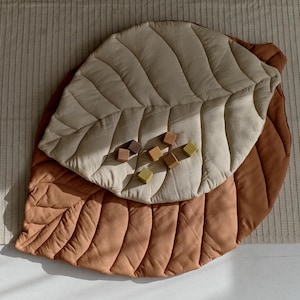 May include: A large, quilted leaf-shaped play mat in brown and beige with a textured surface. The mat is made of two layers of fabric, with the top layer being beige and the bottom layer being brown. There are wooden blocks scattered on the mat.