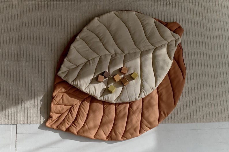 May include: A large, quilted leaf-shaped play mat in brown and beige with a textured surface. The mat is made of two layers of fabric, with the top layer being beige and the bottom layer being brown. There are wooden blocks scattered on the mat.