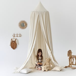 Puede incluir: Una tienda de campaña de algodón blanco para el área de juegos de un niño. Una niña está sentada dentro de la tienda con un oso de peluche y un carro de juguete. Hay un caballo de balancín y un espejo en la pared detrás de la tienda.
