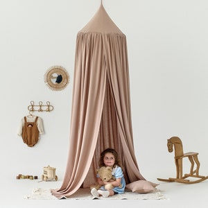 Può includere: Una tenda a baldacchino di colore marrone chiaro con una bambina seduta all'interno che tiene un orsacchiotto. La tenda è installata in una stanza con una parete bianca e un cavallo a dondolo di legno.
