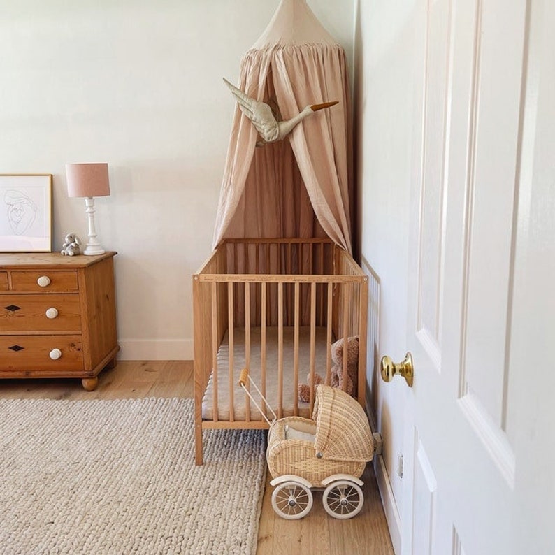 May include: A wooden crib with a white canopy and a stuffed bird hanging from the top. A doll stroller is sitting in front of the crib. The crib is in a room with a wooden dresser and a rug on the floor.