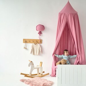 May include: A pink canopy tent hangs over a white crib with a child sitting inside. The crib has a white wooden frame and a white mattress. The child is holding a teddy bear. There is a rocking horse in the foreground.