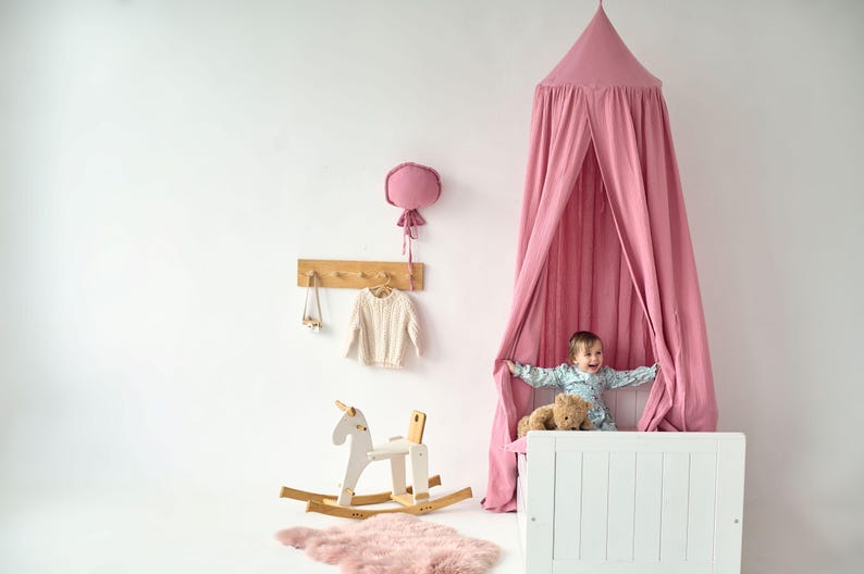 May include: A pink canopy tent hangs over a white crib with a child sitting inside. The crib has a white wooden frame and a white mattress. The child is holding a teddy bear. There is a rocking horse in the foreground.