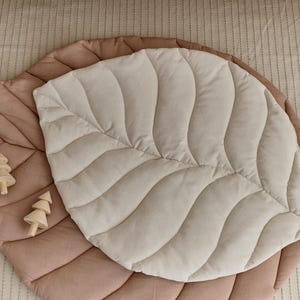 May include: A large, quilted leaf-shaped play mat in beige and brown. The mat is made of soft, plush fabric and has a quilted design. Two small wooden toy trees are placed on the mat.