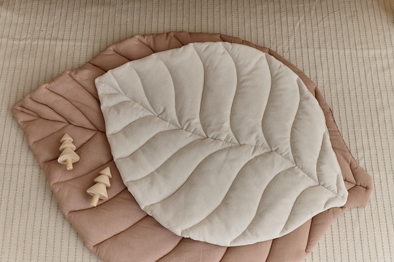 May include: A large, quilted leaf-shaped play mat in beige and brown. The mat is made of soft, plush fabric and has a quilted design. Two small wooden toy trees are placed on the mat.