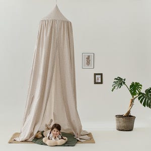 Pu&ograve; includere: Una tenda a baldacchino a quadri beige e marrone con un bambino sdraiato all'interno. La tenda &egrave; realizzata in un tessuto morbido e ha una parte superiore arricciata. Il bambino &egrave; sdraiato su un tappeto verde e marrone con un peluche.