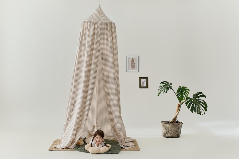 Pu&ograve; includere: Una tenda a baldacchino a quadri beige e marrone con un bambino sdraiato all'interno. La tenda &egrave; realizzata in un tessuto morbido e ha una parte superiore arricciata. Il bambino &egrave; sdraiato su un tappeto verde e marrone con un peluche.
