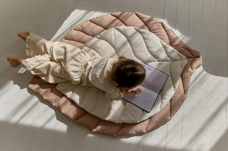May include: A child sits on a large, quilted leaf-shaped play mat in shades of beige and pink. The mat has a quilted design and is perfect for tummy time or playtime. The child is holding a notebook and is drawing or writing.