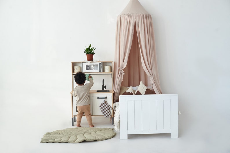 May include: A child plays in a white play kitchen with a green leaf-shaped play mat. A white bed with a pink canopy is in the background.