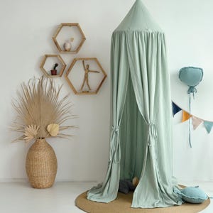 May include: A light green canopy tent with a white background. The tent is made of a soft, flowing fabric and has a gathered top. There are wooden shelves with geometric shapes on the wall behind the tent. A decorative string of flags hangs from the wall.