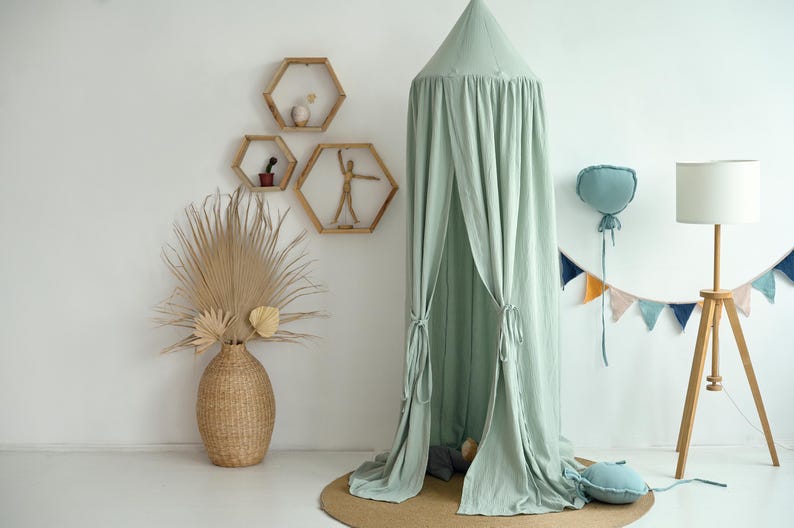 May include: A light green canopy tent with a white background. The tent is made of a soft, flowing fabric and has a gathered top. There are wooden shelves with geometric shapes on the wall behind the tent. A decorative string of flags hangs from the wall.