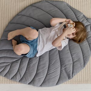 May include: A gray leaf-shaped play mat with a child lying on it, holding a toy camera. The mat has a quilted design with raised lines.
