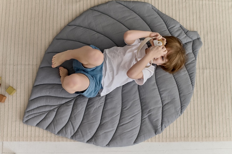 May include: A gray leaf-shaped play mat with a child lying on it, holding a toy camera. The mat has a quilted design with raised lines.