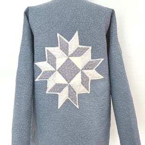 May include: A blue quilted jacket with a large white and gray star-shaped patch on the back. The star is made of fabric with a floral pattern.