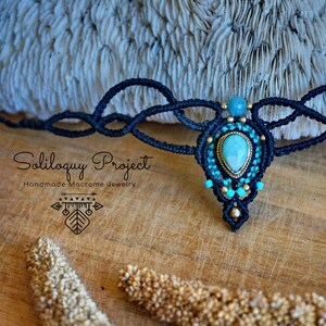 May include: Black macrame choker necklace with a turquoise gemstone pendant. The necklace features a delicate, intricate design and is accented with small gold beads. "Soliloquy Project Handmade Macrame Jewelry" is written on the wooden surface below the necklace.