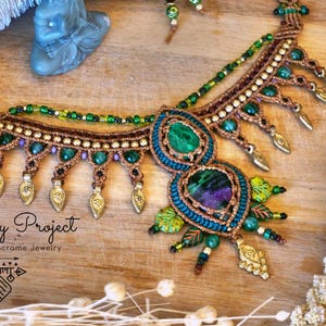 Può includere: Collana macramè fatta a mano con pietre preziose verdi e viola, dettagli color oro e un'intricata lavorazione a nodi. La collana presenta un pendente centrale ed elementi pendenti. Il testo "Soliloquy Project" è visibile.