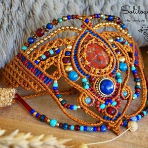 May include: Handmade macrame jewelry piece with intricate details. The piece features a central teardrop-shaped stone, surrounded by blue, red, and gold beads. The macrame is a warm brown color, with the text "Soliloquy Project" visible.