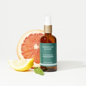 May include: A glass bottle of Moroccan Elixir Slenderize Detoxifying Body Oil, with a green label and gold cap. The bottle is sitting on a white surface with a grapefruit half and a lemon wedge.