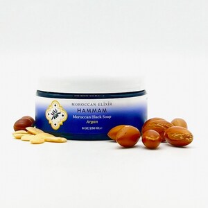 Puede incluir: Un tarro azul de jab&oacute;n negro marroqu&iacute; con aceite de arg&aacute;n. El tarro est&aacute; etiquetado con "Moroccan Elixir Hammam Moroccan Black Soap Argan 8 OZ/250 ML". Hay varias nueces de arg&aacute;n esparcidas alrededor del tarro.