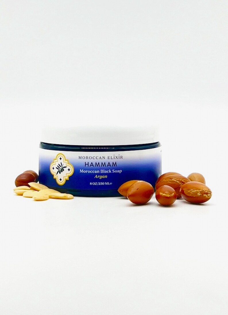 Puede incluir: Un tarro azul de jab&oacute;n negro marroqu&iacute; con aceite de arg&aacute;n. El tarro est&aacute; etiquetado con "Moroccan Elixir Hammam Moroccan Black Soap Argan 8 OZ/250 ML". Hay varias nueces de arg&aacute;n esparcidas alrededor del tarro.