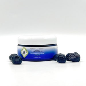 Puede incluir: Un tarro blanco con tapa azul y etiqueta. La etiqueta dice "Moroccan Elixir Hammam Jab&oacute;n negro marroqu&iacute; Natural 8 OZ/230 ML."