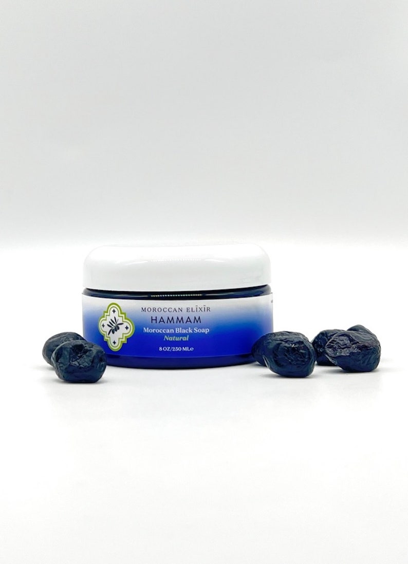 Puede incluir: Un tarro blanco con tapa azul y etiqueta. La etiqueta dice "Moroccan Elixir Hammam Jab&oacute;n negro marroqu&iacute; Natural 8 OZ/230 ML."