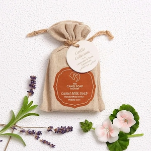 May include: A small, beige cloth bag with a drawstring closure contains a bar of camel milk soap. The soap is labelled "Camel Milk Soap" and "Handcrafted in the Middle East." A white circular tag attached to the bag reads "Castile Collection Lavender & Rose Geranium" in English and Arabic.