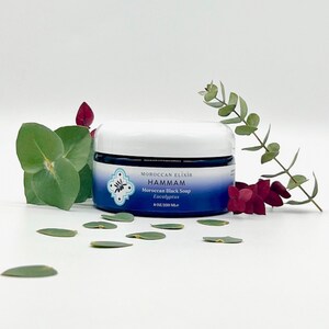Puede incluir: Un tarro azul y blanco de jab&oacute;n negro marroqu&iacute; con aroma a eucalipto de Moroccan Elixir Hammam. El tarro es de 8 oz (250 ml) y est&aacute; sobre una superficie blanca con hojas verdes esparcidas a su alrededor.