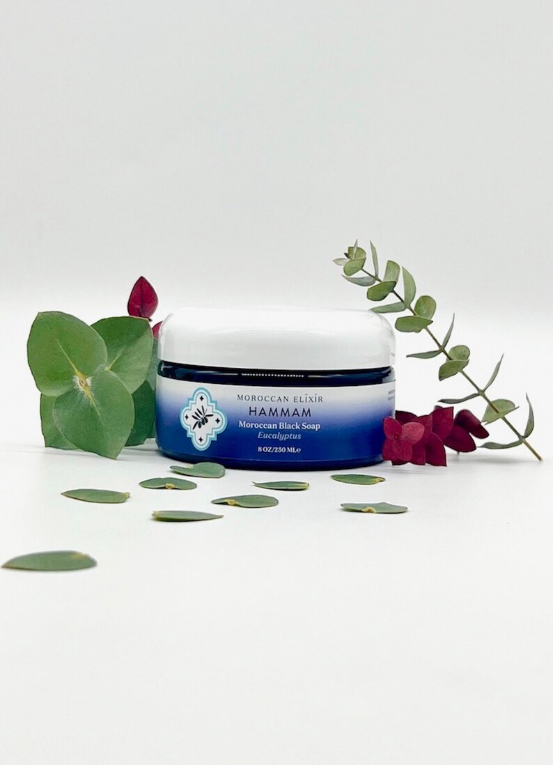 Puede incluir: Un tarro azul y blanco de jab&oacute;n negro marroqu&iacute; con aroma a eucalipto de Moroccan Elixir Hammam. El tarro es de 8 oz (250 ml) y est&aacute; sobre una superficie blanca con hojas verdes esparcidas a su alrededor.