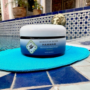 Puede incluir: Un tarro blanco de jab&oacute;n negro marroqu&iacute; natural Hammam Moroccan Elixir, 8 oz (236 ml), sobre un puff de ba&ntilde;o azul.