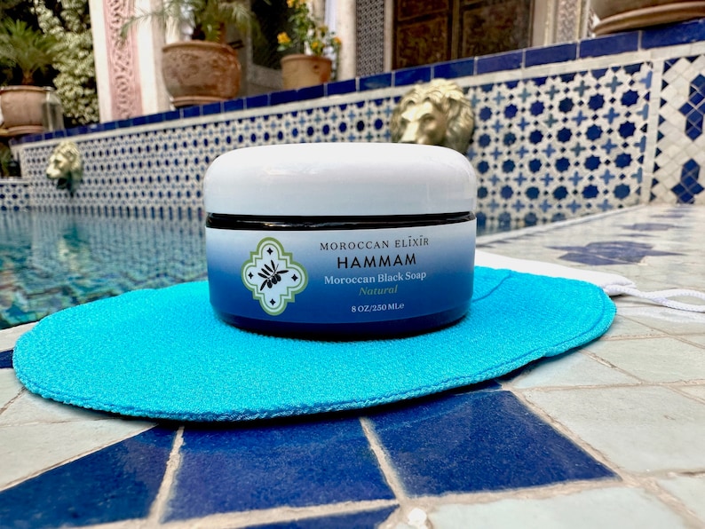 Puede incluir: Un tarro blanco de jab&oacute;n negro marroqu&iacute; natural Hammam Moroccan Elixir, 8 oz (236 ml), sobre un puff de ba&ntilde;o azul.