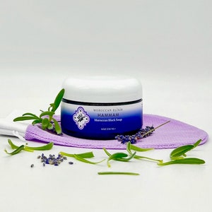 Puede incluir: Un tarro blanco con una etiqueta azul que dice "Moroccan Elixir Hammam Jab&oacute;n Negro Marroqu&iacute; 8 oz/230 ml" est&aacute; sentado en una toallita de ba&ntilde;o morada con ramitas verdes y flores de lavanda esparcidas a su alrededor.