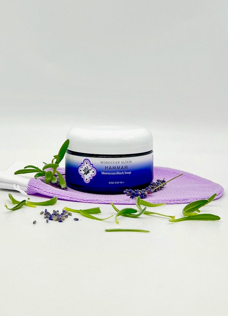 Puede incluir: Un tarro blanco con una etiqueta azul que dice "Moroccan Elixir Hammam Jab&oacute;n Negro Marroqu&iacute; 8 oz/230 ml" est&aacute; sentado en una toallita de ba&ntilde;o morada con ramitas verdes y flores de lavanda esparcidas a su alrededor.