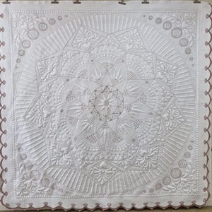 May include: A white quilt with intricate, geometric patterns and floral designs. The quilt features a central starburst motif surrounded by radiating lines and detailed embroidery. The edges are finished with a scalloped border in a muted mauve color.