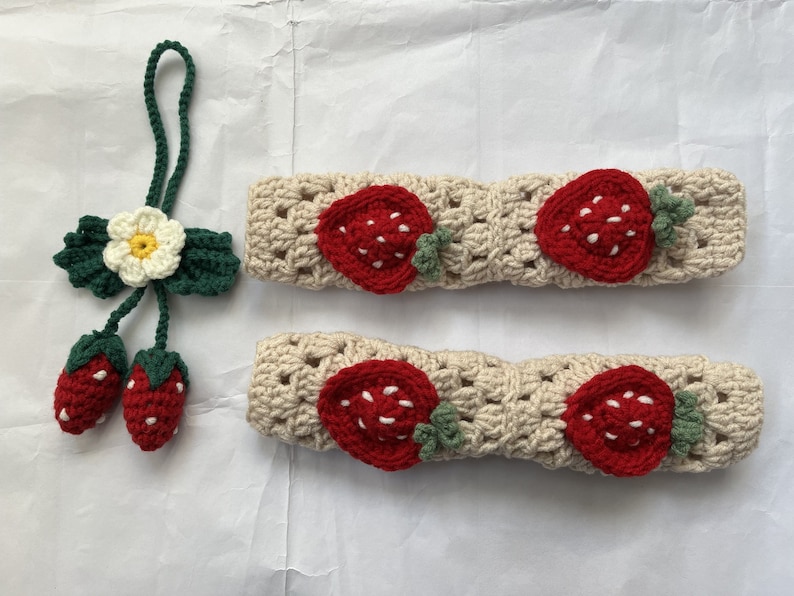 May include: Two beige crocheted headbands with strawberry designs. Each headband has three red strawberries with green leaves.
