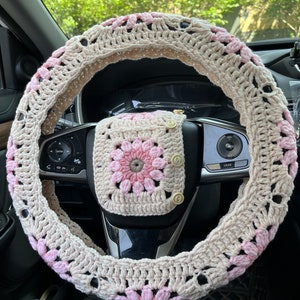 K&ouml;nnte beinhalten: Ein geh&auml;kelter Lenkradbezug in Beige und Rosa mit einem passenden geh&auml;kelten Quadrat mit Kn&ouml;pfen. Der Bezug ist mit einem Granny-Square-Muster gearbeitet.