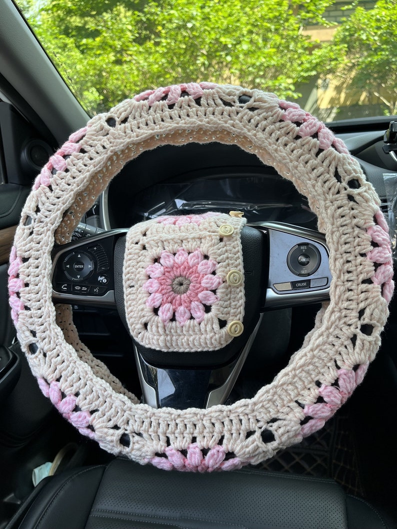 K&ouml;nnte beinhalten: Ein geh&auml;kelter Lenkradbezug in Beige und Rosa mit einem passenden geh&auml;kelten Quadrat mit Kn&ouml;pfen. Der Bezug ist mit einem Granny-Square-Muster gearbeitet.