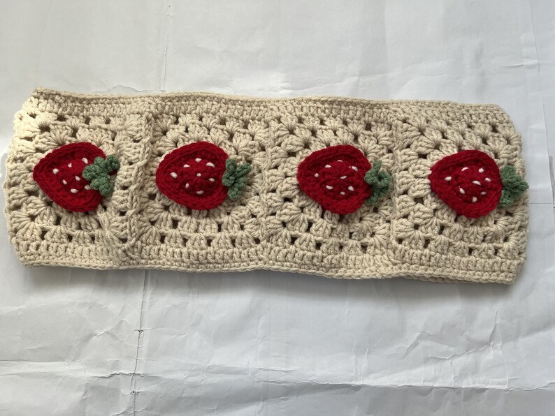 May include: A crocheted headband with a beige background and four red strawberry appliques. Each strawberry has green leaves.