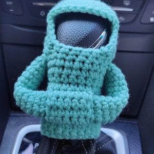 May include: A teal green crocheted  gear shift cover shaped like a person with a hood. The cover is on a black gear shift lever in a car.