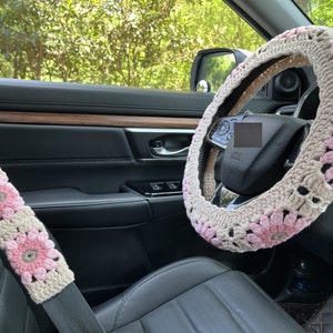 K&ouml;nnte beinhalten: Ein rosafarbener und wei&szlig;er geh&auml;kelter Lenkradbezug mit Blumenmuster. Der Bezug ist auf einem schwarzen Lederlenkrad in einem Auto angebracht.