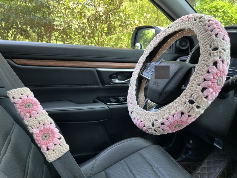K&ouml;nnte beinhalten: Ein rosafarbener und wei&szlig;er geh&auml;kelter Lenkradbezug mit Blumenmuster. Der Bezug ist auf einem schwarzen Lederlenkrad in einem Auto angebracht.