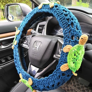 May include: A blue crocheted steering wheel cover with a crocheted turtle design. The cover is decorated with multiple turtles in shades of green, brown, and beige.