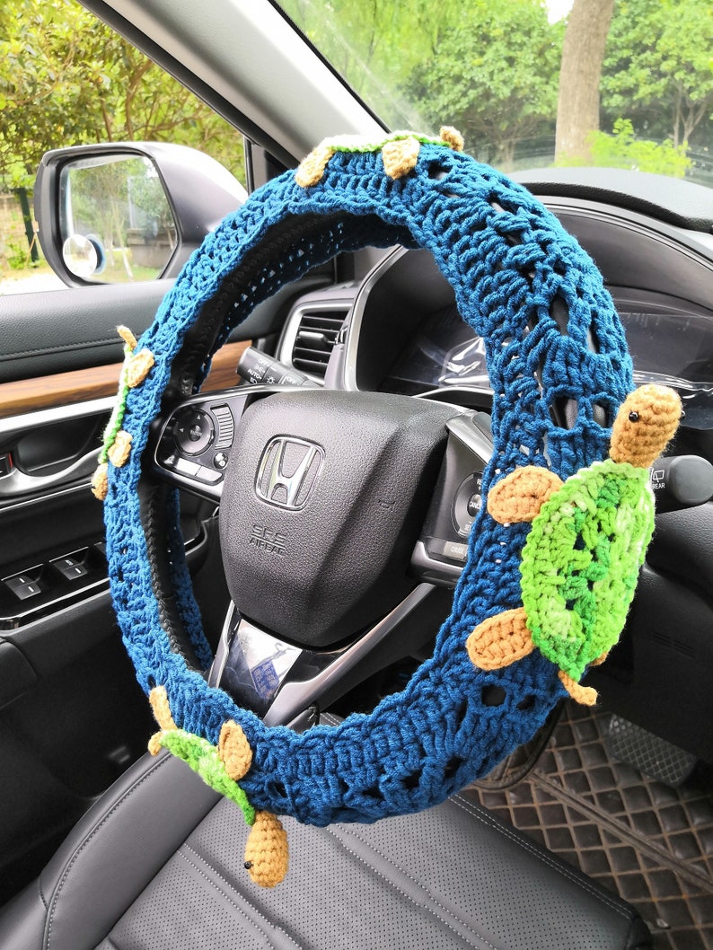 May include: A blue crocheted steering wheel cover with a crocheted turtle design. The cover is decorated with multiple turtles in shades of green, brown, and beige.