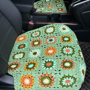 Pu&ograve; includere: Una fodera per sedile auto in crochet verde con un motivo floreale in tonalit&agrave; arancio, giallo, verde e blu.