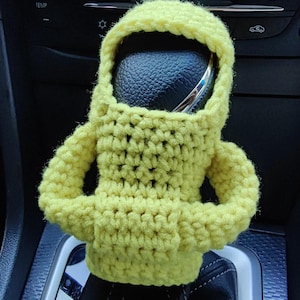 May include: A yellow crocheted gear stick cover shaped like an astronaut. The cover is made of soft yarn and has a round head with a black leather gear stick knob inside.